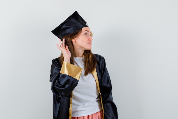 Vrouwelijke afgestudeerde hand achter oor houden in uniform, vrijetijdskleding en nieuwsgierig kijken. vooraanzicht.