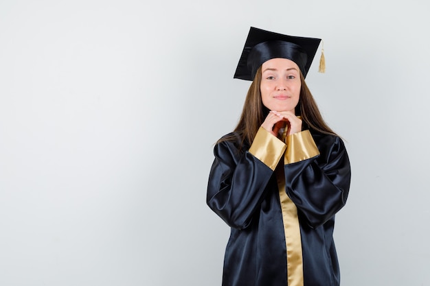 Vrouwelijke afgestudeerde in academische jurk die kin op handen steunt en op zoek dromerig, vooraanzicht.