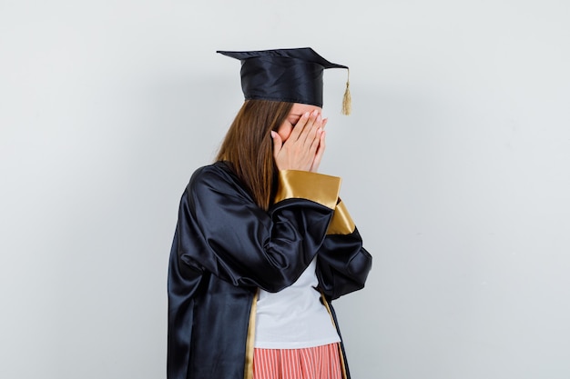 Vrouwelijke afgestudeerde in uniform, vrijetijdskleding die gezicht bedekt met handen en depressief, vooraanzicht kijkt.