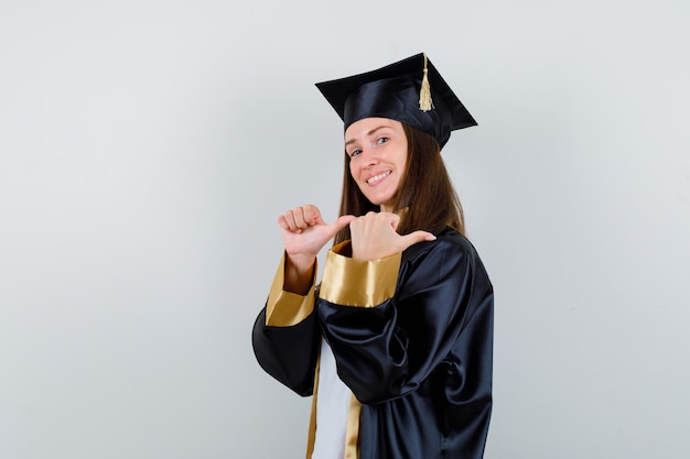 Vrouwelijke afgestudeerde in uniform, vrijetijdskleding wijst met duimen terug en kijkt hoopvol, vooraanzicht.