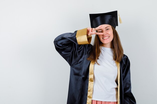 Vrouwelijke afgestudeerde in uniforme, vrijetijdskleding die overwinningsgebaar op oog toont en vrolijk, vooraanzicht kijkt.