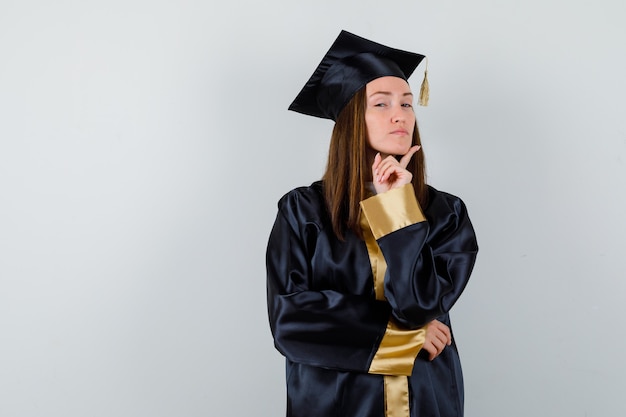 Gratis foto vrouwelijke afgestudeerde propping kin op vinger in academische jurk en op zoek trots, vooraanzicht.