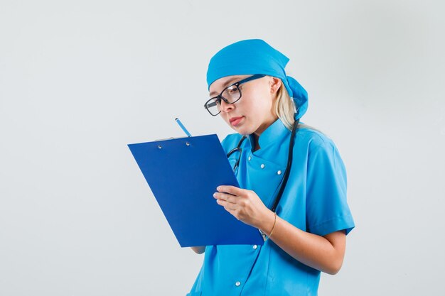 Vrouwelijke arts die aantekeningen maakt op klembord in blauw uniform, glazen en bezig kijkt.