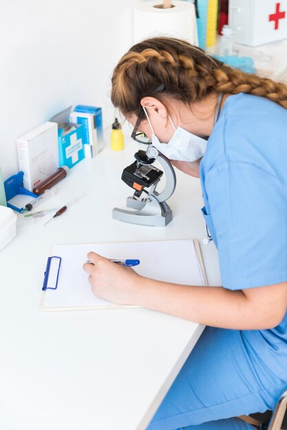 Vrouwelijke arts die door een microscoop kijkt die op klembord in een laboratorium schrijft