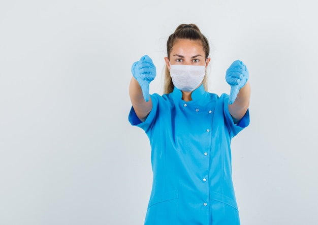 Vrouwelijke arts die duimen neer in blauw uniform, masker, handschoenen toont en ontevreden kijkt.