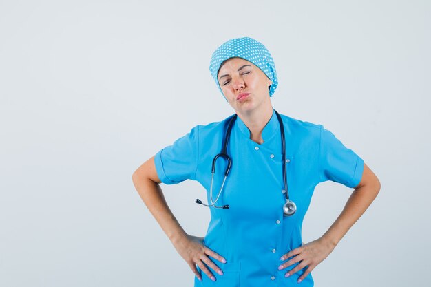 Vrouwelijke arts die lippen pruilde, ogen in blauw uniform sluit en peinzend, vooraanzicht kijkt.