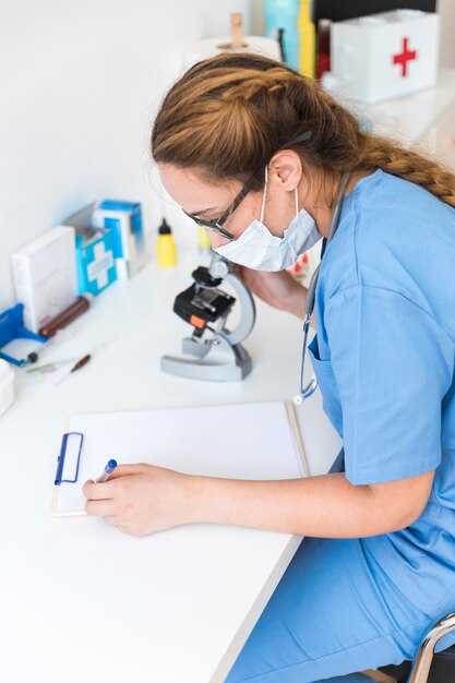 Vrouwelijke arts die masker draagt ​​dat op klembord in een laboratorium schrijft