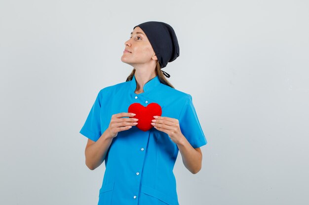 Vrouwelijke arts die rood hart houdt en naar kant kijkt in blauw uniform, zwart hoed vooraanzicht.