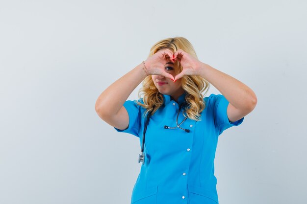 Vrouwelijke arts die vredesgebaar over oog in blauw uniform toont en geliefd kijkt