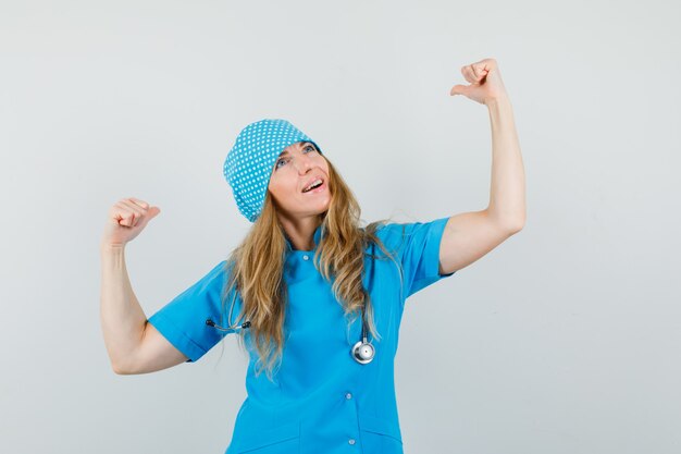 Vrouwelijke arts die winnaargebaar in blauw uniform toont en gelukkig kijkt