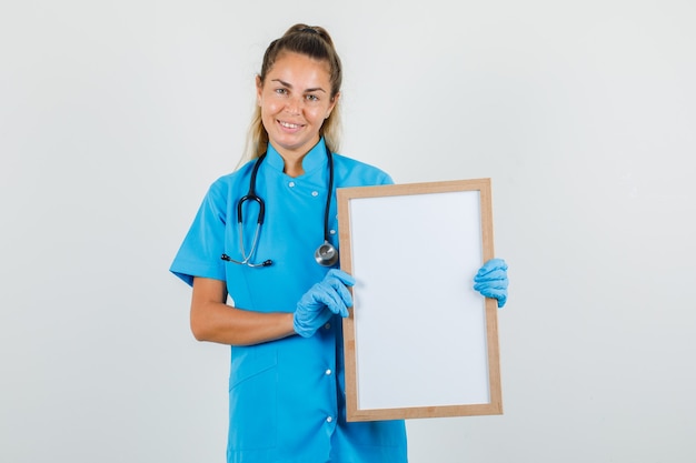 Vrouwelijke arts die wit bord in blauw uniform, handschoenen houdt en vrolijk kijkt.