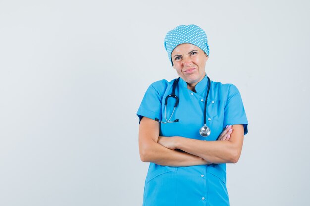 Vrouwelijke arts die zich met gekruiste wapens in blauw uniform bevindt en ontevreden kijkt. vooraanzicht.