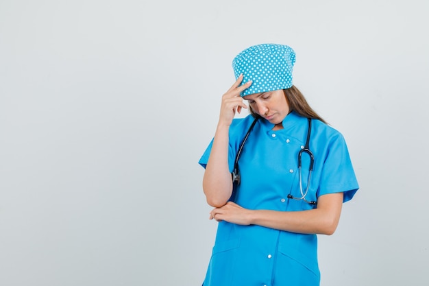 Vrouwelijke arts die zich met hand op hoofd in blauw uniform bevindt en moe kijkt. vooraanzicht.