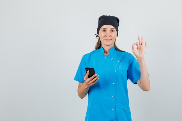 Vrouwelijke arts doet ok teken met smartphone in blauw uniform, zwarte hoed en kijkt tevreden. vooraanzicht.