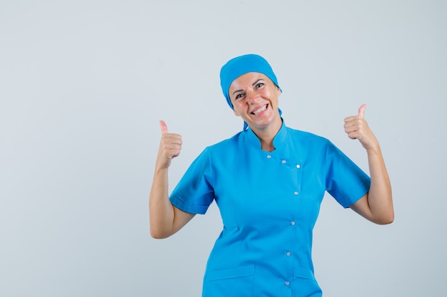 Vrouwelijke arts dubbele duimen opdagen in blauw uniform en op zoek vrolijk, vooraanzicht.