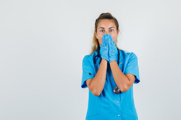 Vrouwelijke arts hand in hand op mond in blauw uniform, handschoenen en op zoek verrast