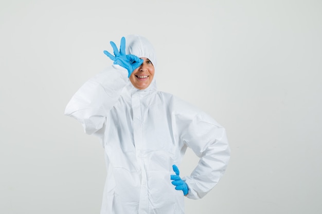 Vrouwelijke arts in beschermend pak, handschoenen die ok teken op oog tonen en grappig kijken