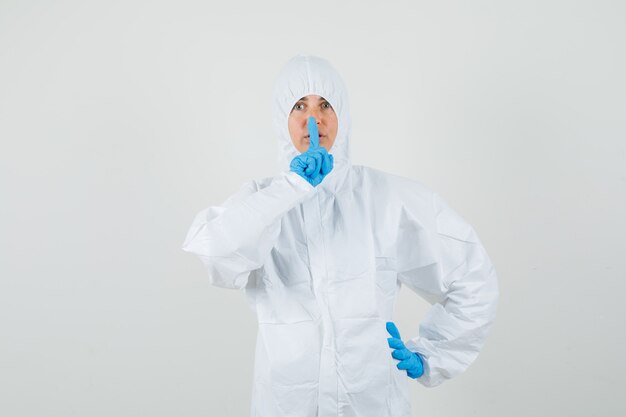 Vrouwelijke arts in beschermend pak, handschoenen die stiltegebaar tonen en voorzichtig kijken
