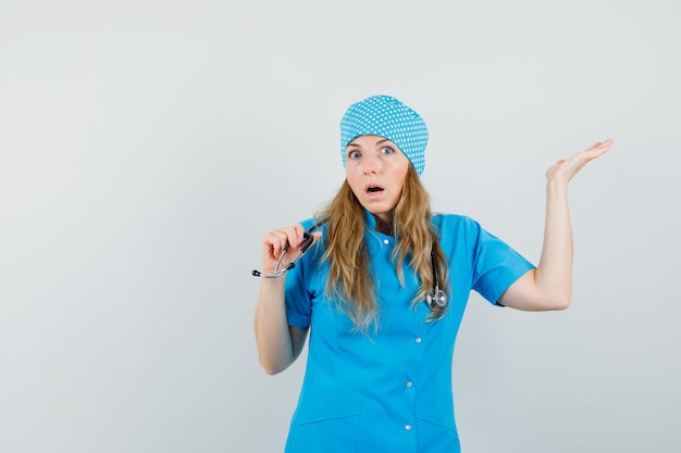 Gratis foto vrouwelijke arts in blauw uniform die stethoscoop houdt terwijl hij palm opheft en verbaasd kijkt