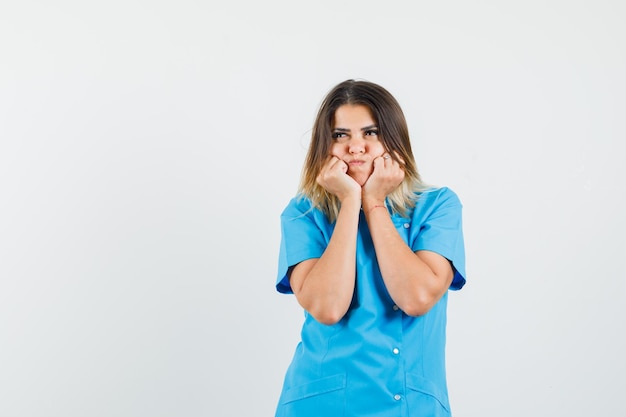 Vrouwelijke arts in blauw uniform die vuisten op de wangen drukt en peinzend kijkt