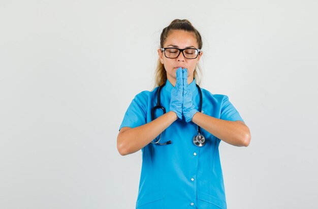 Vrouwelijke arts in blauw uniform, handschoenen, bril hand in hand in biddend gebaar en hoopvol op zoek