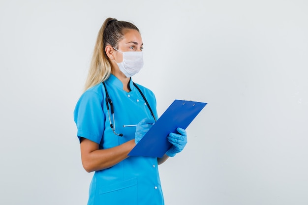 Vrouwelijke arts in blauw uniform, masker, handschoenen die potlood en klembord houden en peinzend kijken