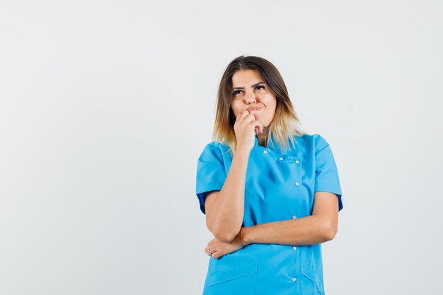 Vrouwelijke arts in blauw uniform staat in denkende houding en kijkt besluiteloos