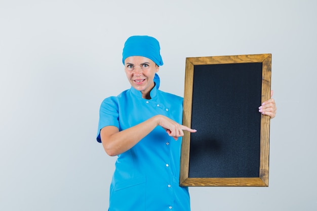 Vrouwelijke arts in blauw uniform wijzend op bord en kijkt vrolijk, vooraanzicht.