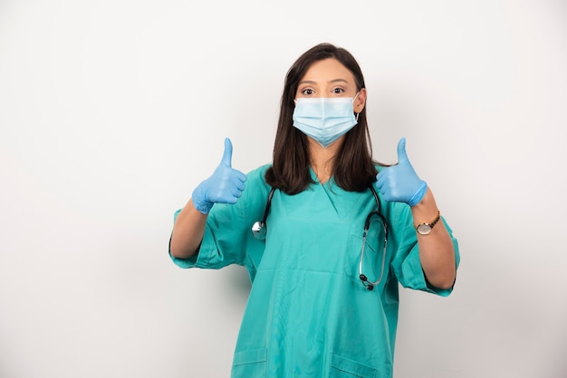 Vrouwelijke arts in masker duimen opdagen op witte achtergrond. Hoge kwaliteit foto