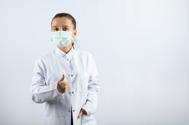 Vrouwelijke arts in wit uniform draagt een medisch masker met een duim omhoog
