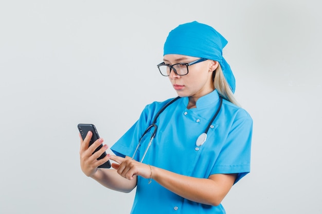 Vrouwelijke arts met behulp van smartphone in blauw uniform, bril en bezig op zoek
