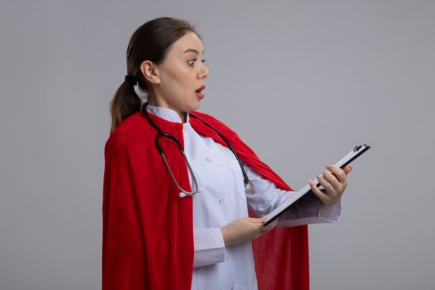Vrouwelijke arts met een stethoscoop in wit medisch uniform en rode superheld cape met klembord met blanco pagina's op zoek verbaasd en verrast staande over witte muur