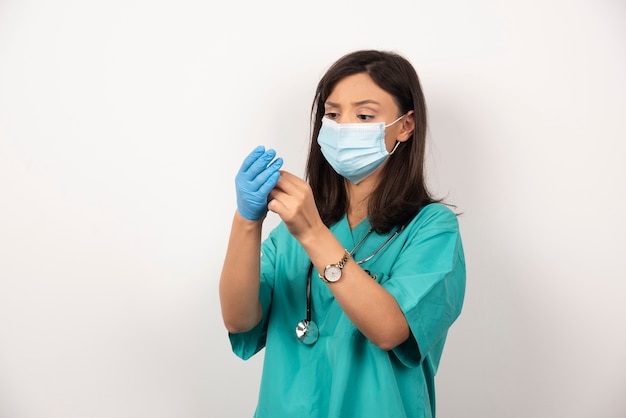 Vrouwelijke arts met medisch masker dat handschoenen op witte achtergrond draagt.