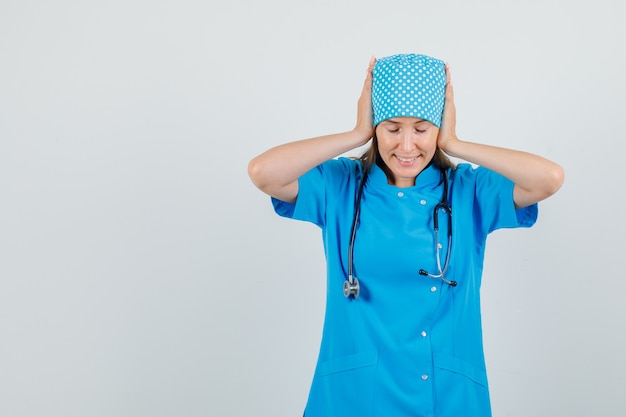 Vrouwelijke arts neerkijkt met de handen op de oren in blauw uniform en kijkt hoopvol. vooraanzicht.