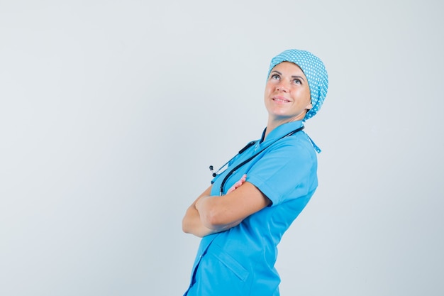 Vrouwelijke arts permanent met gekruiste armen in blauw uniform en peinzend op zoek. .