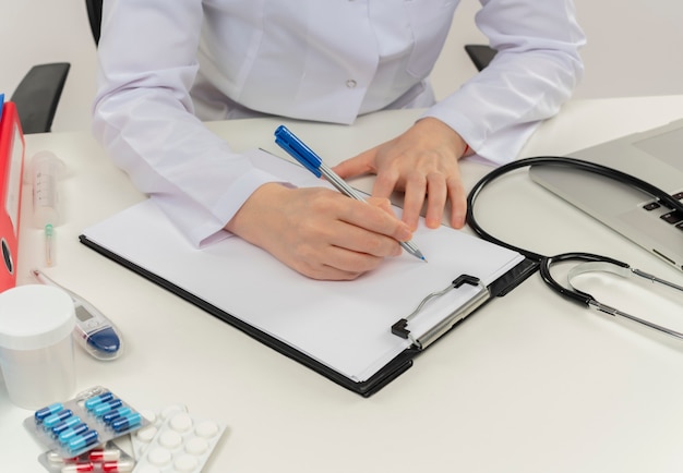 Vrouwelijke arts van middelbare leeftijd die het dragen van medische mantel met een stethoscoop zit aan het bureau werkt op laptop met medische hulpmiddelen schrijft op klembord