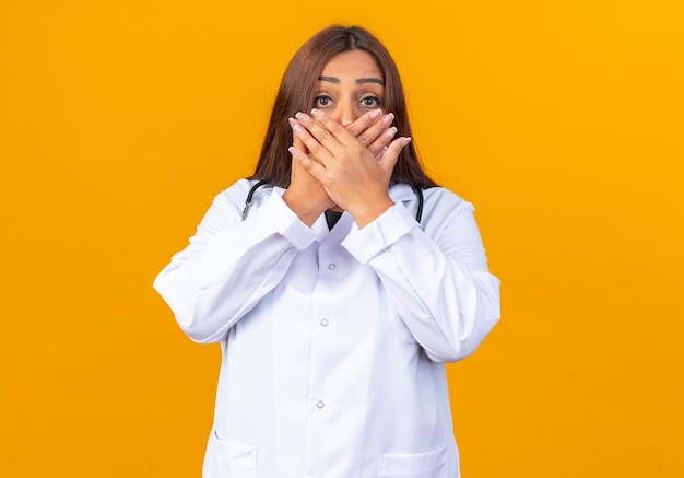 Vrouwelijke arts van middelbare leeftijd in witte jas met stethoscoop die geschokt is over de mond met handen die over oranje muur staan