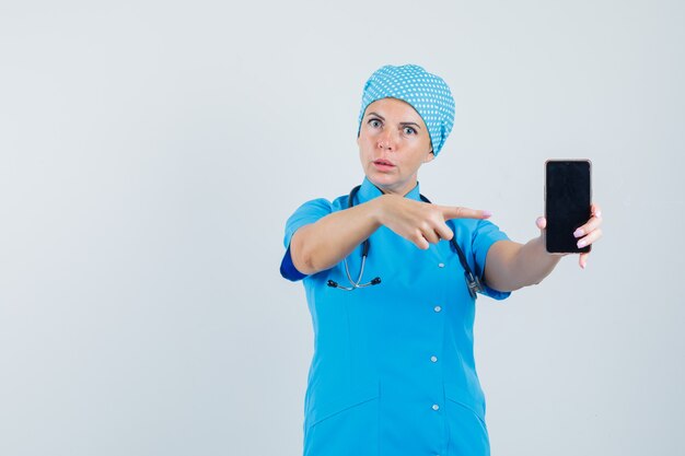 Vrouwelijke arts wijzend op mobiele telefoon in blauw uniform en kijkt zelfverzekerd, vooraanzicht.