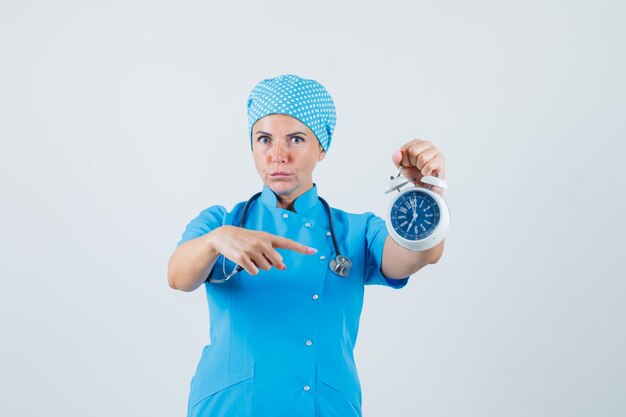 Vrouwelijke arts wijzend op wekker in blauw uniform en op zoek naar ernstige, vooraanzicht.