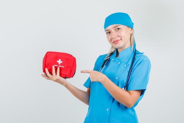 Vrouwelijke arts wijzende vinger op EHBO-kit in blauw uniform en op zoek vrolijk.