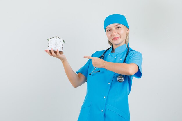 Vrouwelijke arts wijzende vinger op huismodel in blauw uniform en op zoek vrolijk.