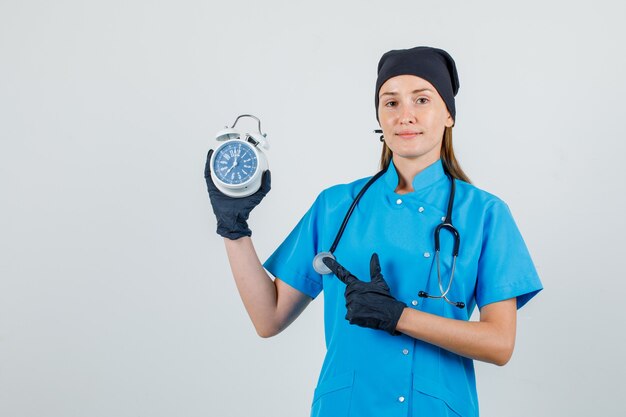 Vrouwelijke arts wijzende vinger op wekker in uniform, handschoenen