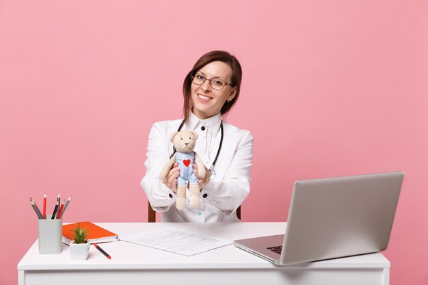 Vrouwelijke arts zit aan bureau werk op computer met medisch document houd speelgoed in ziekenhuis geïsoleerd op pastel roze muur achtergrond. vrouw in medische toga glazen stethoscoop. gezondheidszorg geneeskunde concept.
