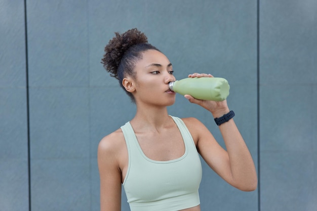 Vrouwelijke atleet met gemotiveerde uitdrukking heeft een trainingspauze en drinkt koud water uit een fles, gekleed in bijgesneden topposities tegen een grijze muur