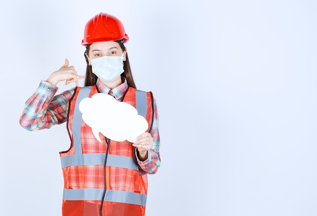 Vrouwelijke bouwingenieur in veiligheidsmasker en rode helm die een infobord in de vorm van een wolk vasthoudt en om een oproep vraagt