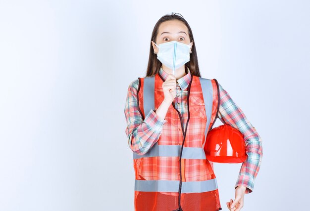 Vrouwelijke bouwingenieur met veiligheidsmasker en een rode helm onder haar armen die een pen vasthoudt en er verward en attent uitziet