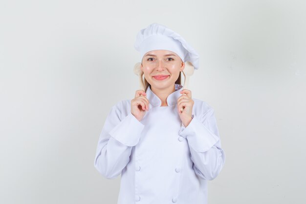 Vrouwelijke chef-kok die houten lepels achter oren in wit uniform houdt en vrolijk kijkt