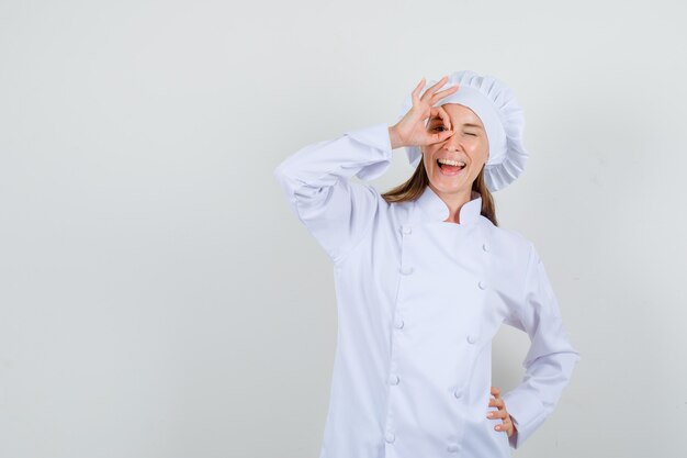 Vrouwelijke chef-kok die ok gebaar op oog in wit uniform toont en optimistisch kijkt. vooraanzicht.