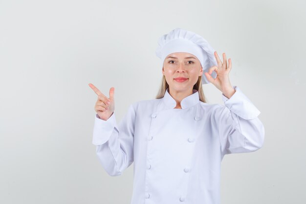 Vrouwelijke chef-kok die ok teken en geweergebaar in wit uniform toont en vrolijk kijkt.