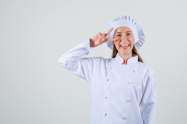 Vrouwelijke chef-kok die overwinningsgebaar dichtbij oog in wit uniform toont en blij, vooraanzicht kijkt.